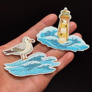 May include: Two embroidered patches featuring nautical themes. One patch depicts a white and grey seagull standing on blue ocean waves. The second patch shows a yellow and white lighthouse surrounded by stylised blue ocean waves and sea foam.