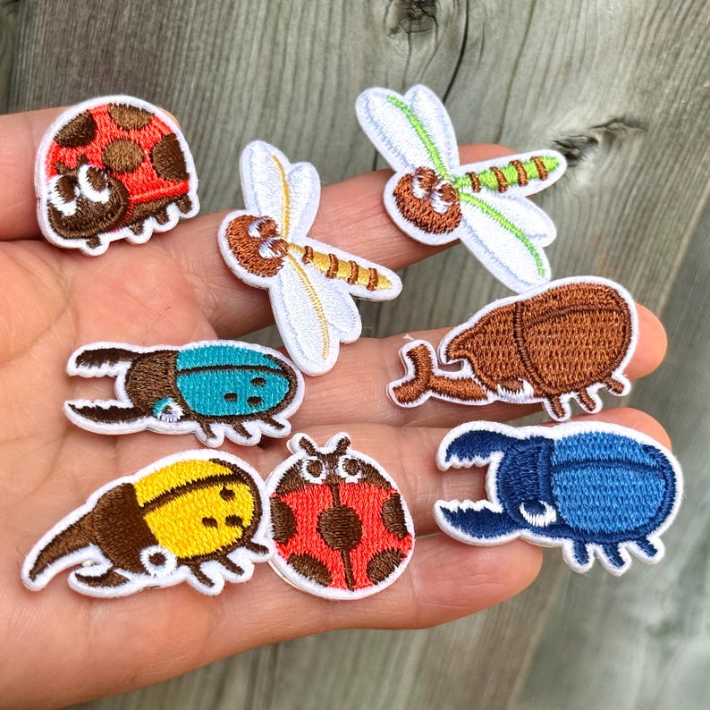 May include: A selection of embroidered insect patches, including ladybirds, dragonflies, and beetles. The patches feature various colours such as red, blue, yellow, and brown, with detailed stitching and white borders. These patches are ideal for adding to clothing or accessories.