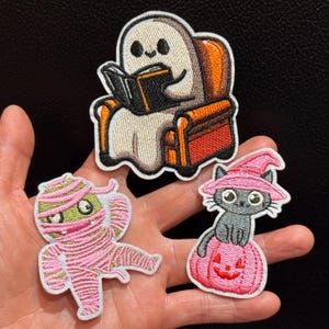 Puede incluir: Tres parches bordados con temas de Halloween: un fantasma blanco leyendo un libro negro en un sillón naranja, una momia verde envuelta en vendas rosas y un gato gris con un sombrero de bruja rosa sobre una calabaza rosa.