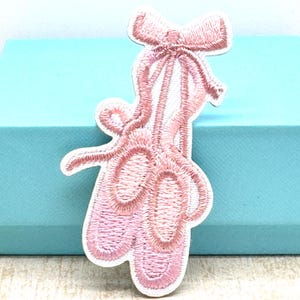 May include: Embroidered patch of pink ballet pointe shoes with a bow. The patch has a white border and is designed for application on fabric items. The design is detailed and delicate, suitable for adding to clothing or accessories.