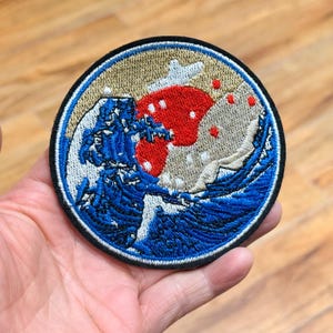 May include: Embroidered patch with a circular design depicting a wave and red and white elements. The patch has a blue border and a black edge. The wave is in shades of blue and white, with a beige background.