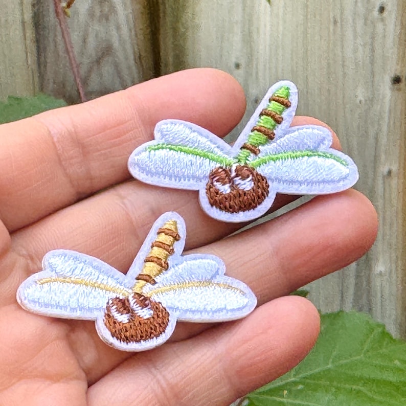May include: Two embroidered dragonfly patches. Each patch features white wings with green trim, a brown body, and a segmented stem in shades of brown and green. The patches are small and detailed.