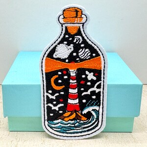 May include: Embroidered patch in the shape of a bottle, featuring a lighthouse scene. The design includes a red and white striped lighthouse, waves, birds, a crescent moon, stars, and planets against a black background. The patch has a white border.