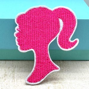 May include: Pink embroidered iron-on patch of a woman's silhouette with a ponytail.