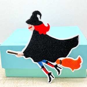 May include: Embroidered patch of a witch on a broomstick. The witch is depicted with a black hat and cloak, red hair, and a smiling face. Her legs are in blue trousers and red-striped boots. The broom has an orange brush.