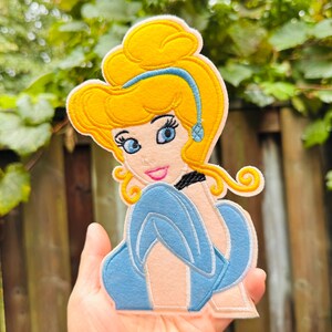 Pode incluir: Patch bordado com uma imagem de desenho animado da Cinderela. O patch tem um penteado amarelo, olhos azuis e um vestido azul claro. O patch é feito de feltro e tem um fundo rosa.