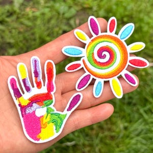 May include: Two embroidered patches featuring vibrant, multi-coloured designs. One patch is shaped like a handprint with rainbow-coloured segments, and the other is a circular sun motif with a central spiral and radiating petals in various bright hues.
