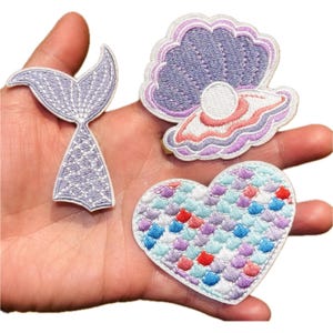 Puede incluir: Tres parches bordados: una cola de sirena, una concha con una perla y un parche en forma de corazón con un patrón de escamas. La cola de sirena es lavanda con costuras blancas. La concha es morada, rosa y blanca. El corazón es azul, rojo y morado.
