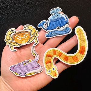 Peut inclure: Écussons brodés représentant des créatures marines : un crabe, une baleine, une raie et un ver. Les écussons présentent des coutures détaillées et des couleurs vives, notamment le jaune, le bleu, le violet et l'orange. Chaque écusson a une bordure blanche.