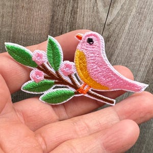 May include: Embroidered patch of a pink bird with a yellow chest and orange beak, sitting on a branch with green leaves and pink flowers. The patch has a white border, suitable for clothing or accessories.