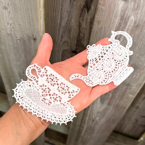 May include: Two white lace appliqués shaped like a teapot and a teacup with a saucer. The intricate needlework features delicate floral and geometric patterns with scalloped edges, crafted from fine white thread in a detailed openwork design.