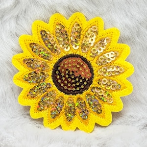 May include: A yellow felt sunflower with a brown centre and gold sequins. The petals are outlined with yellow stitching. The centre of the flower is a dark brown colour with small gold sequins. The sunflower is a decorative item.