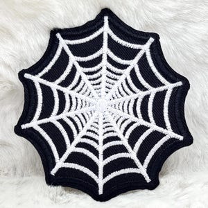 以下が含まれることがあります： 白と黒の刺繍されたクモの巣のパッチ。パッチは、黒い背景に詳細な白いウェブデザインが特徴です。ウェブは中央に配置され、外側に放射状に広がり、対称的なパターンを作成します。パッチはおそらく装飾目的です。
