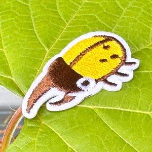 May include: Embroidered patch of a yellow banana with brown details and white edging. The patch is set against a green leaf. The design is a playful, cartoon-style depiction of a banana.