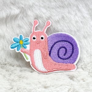 May include: Embroidered patch featuring a smiling pink snail holding a blue flower with a yellow center. The snail has a purple shell with a spiral design and white accents. The patch is designed with a white border.