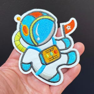 May include: Embroidered patch featuring a cartoon astronaut in a white spacesuit with blue and orange accents. The astronaut holds a flag with a star. The patch has a yellow tube and a control panel on the chest. The patch is held in a hand.