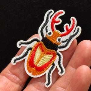 May include: An embroidered patch shaped like a stag beetle with a white border. The beetle features a bright red, orange, and yellow body, a black thorax, and prominent red mandibles. The design includes black legs and antennae on a white background.