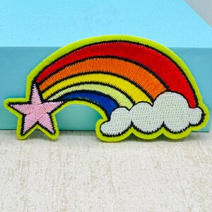 Può includere: Toppa arcobaleno ricamata con una stella rosa e una nuvola bianca. L'arcobaleno presenta strisce rosse, arancioni, gialle e blu, bordate di nero. La toppa ha un bordo verde brillante.