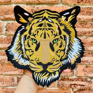 Op de afbeelding: Geborduurde tijgerkop patch met een gedetailleerd ontwerp met gele, zwarte en witte accenten. De tijger heeft groene ogen en een bruine neus. De patch wordt tegen een bakstenen muurachtergrond gehouden.