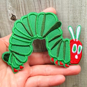 Op de afbeelding: Geborduurde patch met een groene rups met een rode kop en witte antennes. De rups heeft een gesegmenteerd lichaam en rode voeten. De patch is ontworpen met gedetailleerde stiksels, voor kleding of accessoires.
