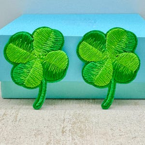 以下が含まれることがあります： 緑色の刺繍された四つ葉のクローバーのパッチが2つ。 クローバーの葉には詳細なステッチが施され、各パッチには緑色の茎があります。 パッチは水色の表面に表示されています。