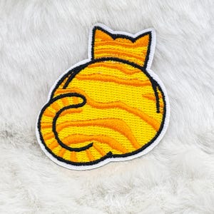 Op de afbeelding: Geborduurde patch met de achterkant van een cartoonkat. De patch heeft een geel en oranje gestreept ontwerp, omlijnd in zwart, met een witte rand. De staart van de kat is gekruld. De patch ligt op een wit, pluizig oppervlak.