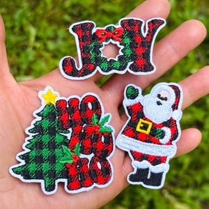 Könnte beinhalten: Drei bestickte Aufnäher mit Weihnachtsmotiven. Ein Aufnäher zeigt das Wort JOY in rot-schwarzem Karomuster mit einem grünen Kranz. Ein weiterer zeigt einen grünen Weihnachtsbaum mit dem Text HO HO HO. Der dritte Aufnäher stellt den Weihnachtsmann dar.