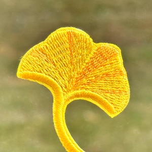 May include: A close-up of a vibrant yellow embroidered ginkgo leaf patch. The patch displays intricate stitching, highlighting the leaf's form and veins. The stem curves elegantly, set against a soft, blurred green backdrop.