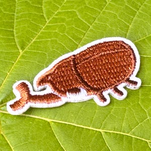 May include: A brown embroidered beetle patch with a white border, set against a green leaf. The patch features detailed stitching, depicting the insect's form. A decorative item.