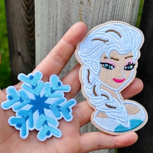 May include: Two embroidered patches: one of a snowflake in shades of blue and white, and another of a cartoon character with light blue hair, pink lips, and blue eyes. The patches are on a beige background.