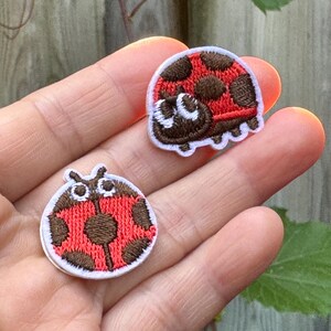 May include: Two embroidered ladybug patches. Each patch features a red body with brown spots and a white border. One ladybug has a smiling face. These patches are ideal for decorating clothing or accessories.