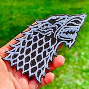Op de afbeelding: Geborduurde patch met een profiel van een direwolf-kop. De patch heeft een zwarte achtergrond met witte stiksels die de kenmerken en vacht van de wolf omlijnen. De patch wordt tegen een groene achtergrond gehouden.