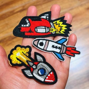 May include: Three embroidered patches featuring space-themed designs. One patch is a red rocket with a yellow exhaust flame, another is a white and blue rocket with red exhaust, and the third is a grey rocket with a large yellow exhaust cloud.