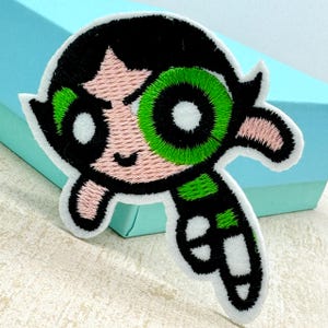 Op de afbeelding: Een geborduurde strijk-op patch met een cartoonfiguur met zwart haar, groene ogen en een roze gezicht. Het personage draagt een groen-zwarte outfit en heeft een ondeugende uitdrukking.