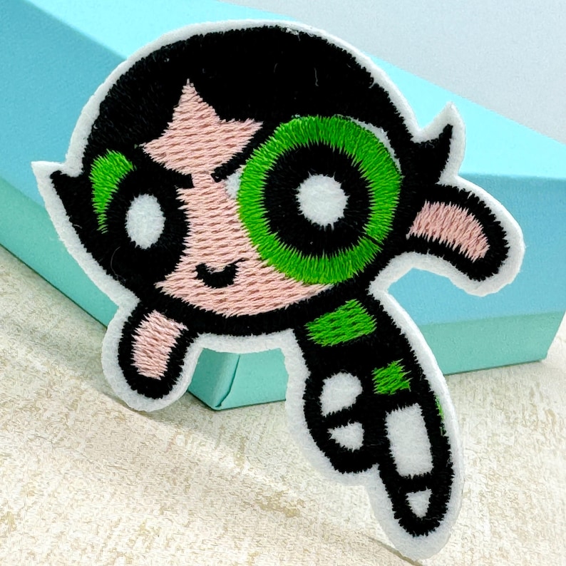 Op de afbeelding: Een geborduurde strijk-op patch met een cartoonfiguur met zwart haar, groene ogen en een roze gezicht. Het personage draagt een groen-zwarte outfit en heeft een ondeugende uitdrukking.