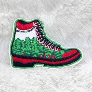 Op de afbeelding: Geborduurde patch in de vorm van een wandelschoen. De schoen heeft een rode zool en accenten, een groen bovenwerk met een boslandschap en zwarte veters. De patch heeft een witte rand.