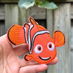 Op de afbeelding: Geborduurde oranje clownvis patch met zwarte, witte en oranje details. De patch toont een lachende vis met grote ogen en een zwarte omtrek. Geschikt voor kleding of accessoires.