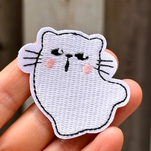 Peut inclure: Un écusson brodé blanc en forme de chat au corps arrondi. Le chat présente des yeux brodés noirs, un petit nez noir et trois moustaches noires de chaque côté. Deux petits cercles roses sont brodés sur les joues pour un effet blush.