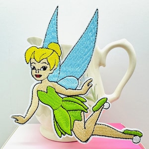 May include: Embroidered iron-on patch featuring Tinker Bell, a fairy with green wings, a green dress, and blonde hair.