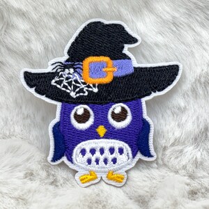 May include: Embroidered patch of a purple owl wearing a black witch's hat with a spider and web. The hat has a purple band with an orange buckle. The owl has large brown eyes, a yellow beak, and yellow feet. The patch is outlined in white.