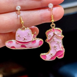 May include: A pair of pink and gold earrings with a pink cowboy hat and a pink cowboy boot design. The earrings have a star pattern.