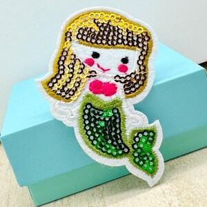 May include: A decorative mermaid patch featuring a white face, pink cheeks, and a pink bow. The mermaid's hair is made of gold sequins, and her green tail is decorated with green sequins. The patch is edged with white stitching.