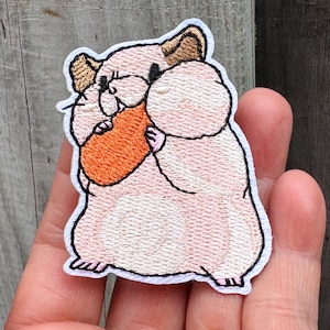 以下が含まれることがあります： ニンジンを持っている漫画のハムスターの刺繍パッチ。 ハムスターは薄いピンク色で、茶色の耳と黒い目があります。 ニンジンはオレンジ色です。 パッチには白い縁があり、衣類やアクセサリー用です。