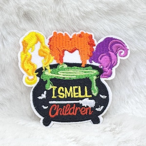 May include: Embroidered patch featuring a black cauldron with green bubbling liquid and three colorful wigs: yellow, orange, and purple. The text "I SMELL Children" is embroidered in green and red. The patch has a white border.