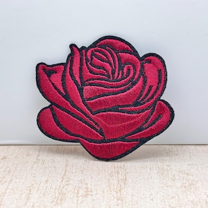 May include: Embroidered red rose patch with a black outline. The patch features detailed stitching, showcasing the petals and overall shape of the flower. This decorative patch can be applied to clothing, bags, or other fabric items.