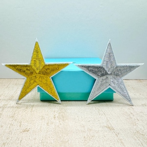 May include: Two star-shaped embroidered patches. One is gold with a white border, the other is silver with a white border. The patches are placed on a light-colored surface with a blue box in the background. These patches are ideal for adding flair to clothing or accessories.