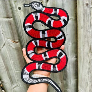 Peut inclure: Écusson brodé en forme de serpent, rouge, noir, blanc et gris. Le motif du serpent présente un corps rouge avec des segments rayés noirs et blancs. La tête du serpent est détaillée avec des motifs gris et blancs, et l'écusson a une bordure noire.