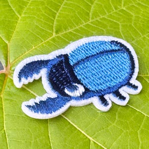 May include: Embroidered patch of a blue beetle with white edging. The patch has a textured finish and is set against a green leaf. The beetle design is detailed and striking, suitable for clothing or accessories.