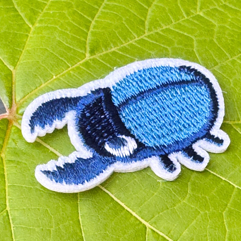 May include: Embroidered patch of a blue beetle with white edging. The patch has a textured finish and is set against a green leaf. The beetle design is detailed and striking, suitable for clothing or accessories.