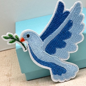May include: Embroidered patch of a blue dove with a green olive branch in its beak. The dove has a white border and is set against a light blue background. The patch is designed for application to clothing or accessories.